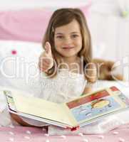 Little girl reading in bed with thumb up