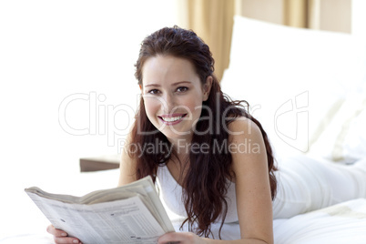 Brunette woman in bed reading a newspaper and smiling
