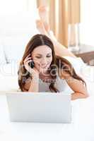 Pretty woman on phone using her laptop lying on bed
