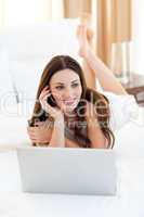 Brunette woman on phone using her laptop lying on bed