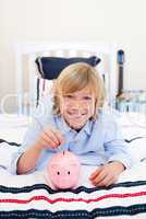 Smiling boy inserting a coin in a piggybank lying down on bed