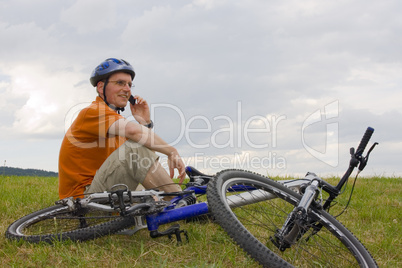 Mann sitzt neben Mountainbike auf Wiese und telefoniert