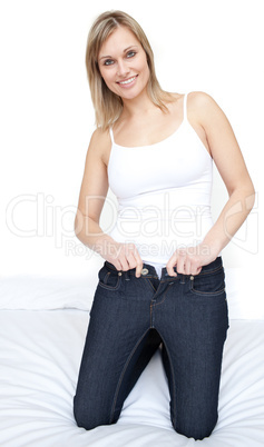 Smiling woman putting on tight jeans on a bed