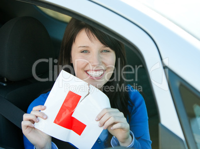 Smiling brunette teen girl sitting in her car tearing a L-sign