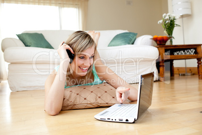 Cheerful woman on phone while using a laptp lying on the floor