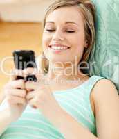 Smiling young woman sending a text lying on a sofa