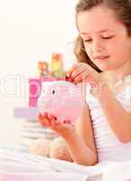 Cute little girl saving money in a piggy-bank sitting on her bed