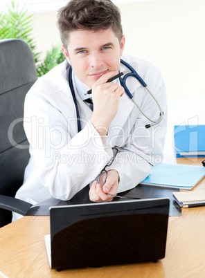 Smiling male doctor using a laptop sitting at his desk