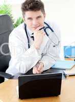 Smiling male doctor using a laptop sitting at his desk