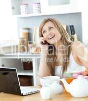 Delighted woman using a phone and laptop in the kitchen