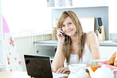 Caucasian woman using a laptop and a phone in the kitchen