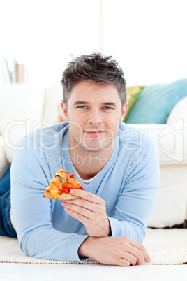 Smiling young man eating a pizza lying on the floor