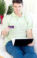 Charismatic young man with card and laptop on a sofa