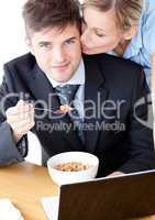 Smiling businessman eating breakfast while wife kissing his chee