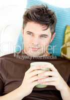 Charismatic young man holding a cup lying on the sofa