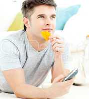 Jolly young man holding a remote eating crisps lying on the fllo