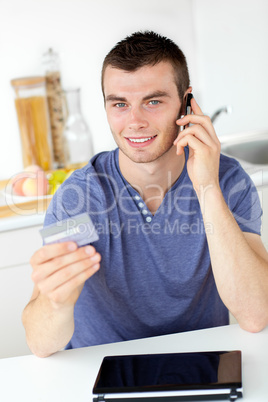 Attractive young man talking on phone holding a card looking at