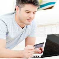 Positive young man holding a card using his laptop on the floor