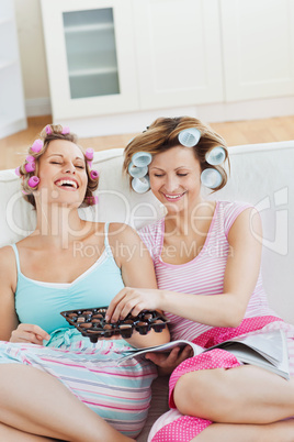 Delighted female friends with hair rollers eating chocolate read