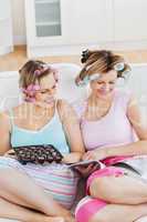 Female friends with hair rollers eating chocolate reading a maga
