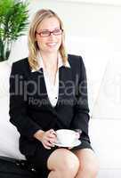 Delighted businesswoman wearing glasses drinking coffee sitting