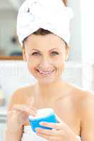Cute young woman with a towel putting cream on her face in the b