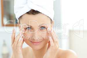 Cute young woman with a towel putting cream on her face in the b