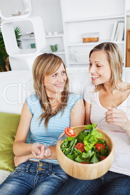 Two female friends eating salad together on a sofa
