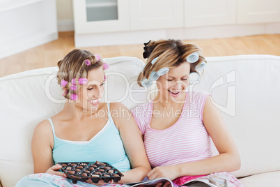 Close female friends with hair rollers eating chocolate reading
