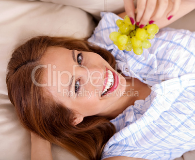 Happy young woman lying on sofa holding grapes