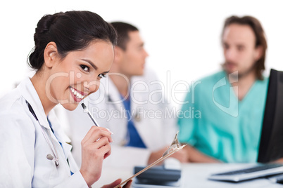 Young asian doctor smiling with the pen and looking the camera, others blurred behind