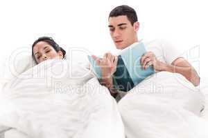 Young handsome man reading book while his wife sleeping beside him in bed