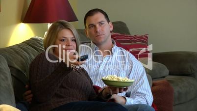 A couple watches television, eating popcorn and fighting over remote.