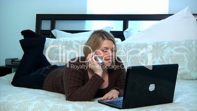 A woman uses her laptop in the bedroom while talking on the phone.