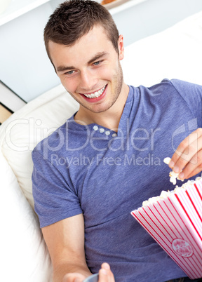 Positive caucasian man holding a remote and eating popcorn in th