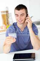 Attractive young man talking on phone holding a card in the kitc