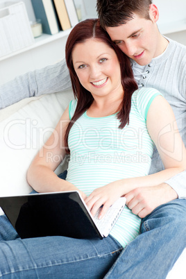 Enthusiastic couple using laptop smiling at the camera sitting o