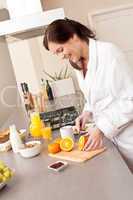 Young woman in bathrobe cutting orange in kitchen