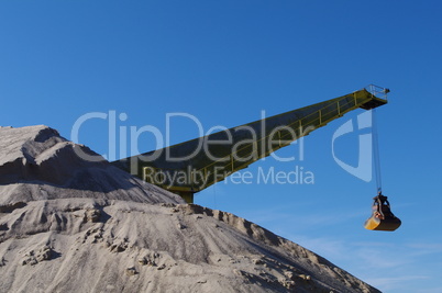 Kran mit gelber Schaufel hinter hohem Sandberg vor strahlend blauem Himmel