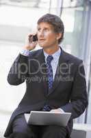 Businessman sitting in office lobby with laptop using cellular p
