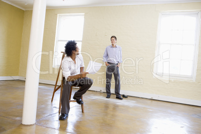 Man sitting on ladder in empty space holding paper talking to ot