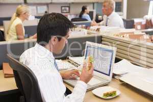 Businessman in cubicle at laptop eating sandwich