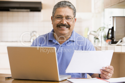 Man in kitchen with laptop and paperwork smiling