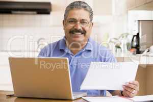 Man in kitchen with laptop and paperwork smiling