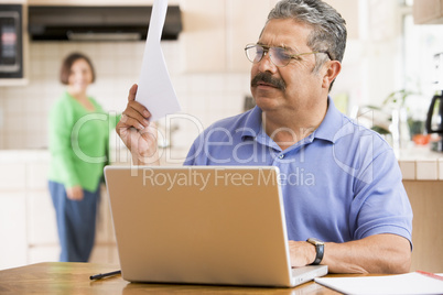 Man in kitchen with laptop and paperwork with woman in backgroun