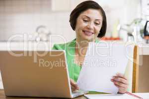 Woman in kitchen with laptop and paperwork smiling