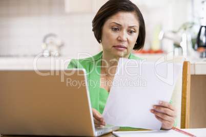 Woman in kitchen with laptop and paperwork looking worried