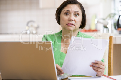 Woman in kitchen with laptop and paperwork looking worried