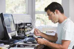 Man in home office using computer holding paperwork and looking