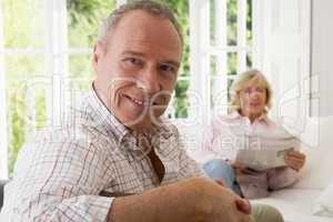 Man in living room smiling with woman in background reading news